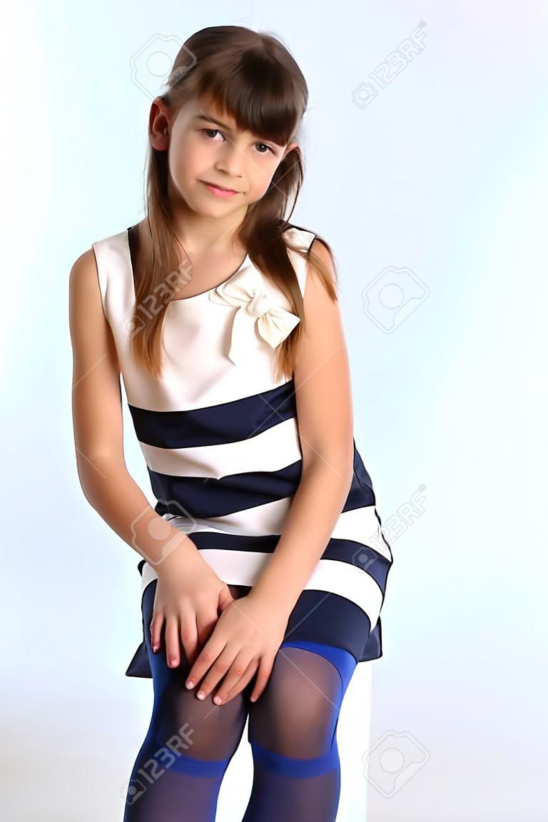 Karcsú, csíkos ruhás gyönyörű lány ült a térdén. Nagyon boldog vonzó gyermek kék harisnya. A fiatal iskoláslány 9 éves.