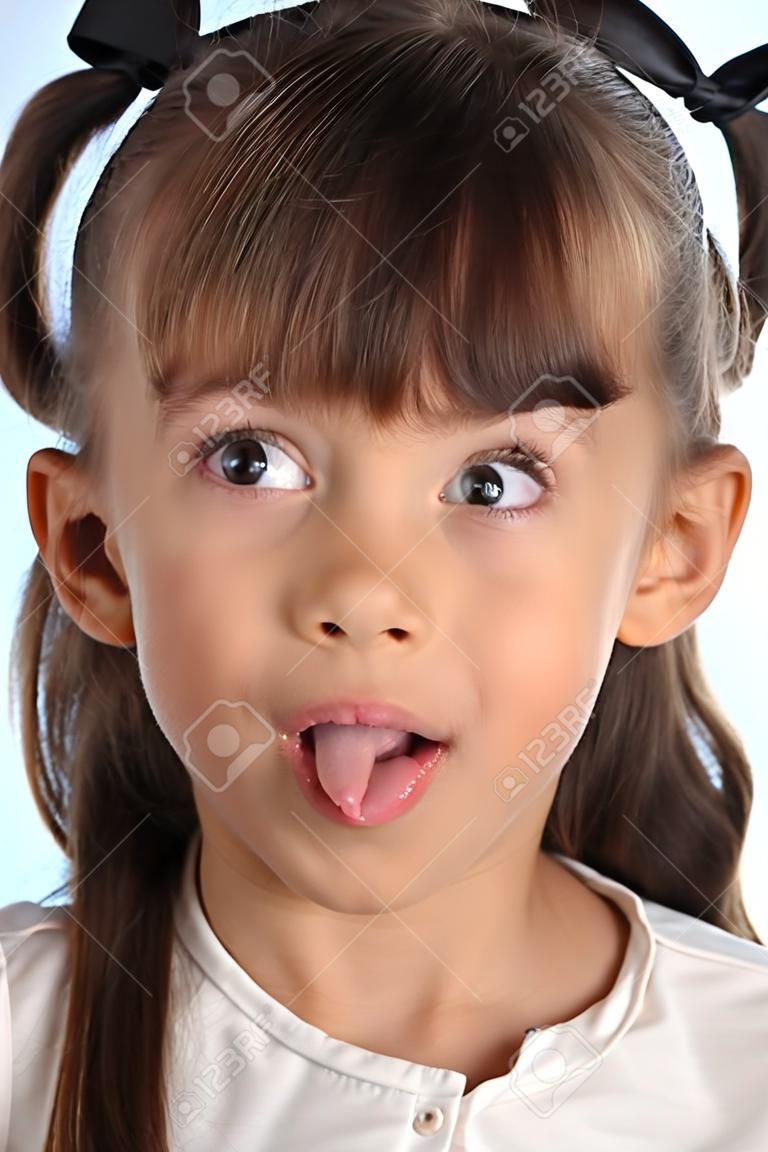 Close-up portrait of a beautiful girl in a white blouse. The cute attractive child teases by pulling her pink tongue and rolling her eyes. The young schoolgirl is 9 years old.