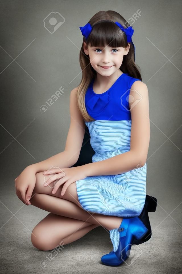 苗条的漂亮女孩，穿着条纹的衣服坐在她的膝盖上。相当蓝色贴身衬衣的愉快的可爱的孩子。年轻的女学生9岁。