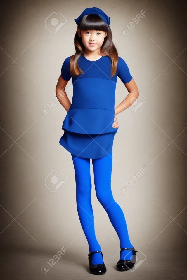 Piękna dziewczyna w pasiastej sukience stoi na całej długości. Eleganckie atrakcyjne dziecko o smukłym ciele i długich nogach w niebieskich rajstopach. Młoda uczennica ma 9 lat.