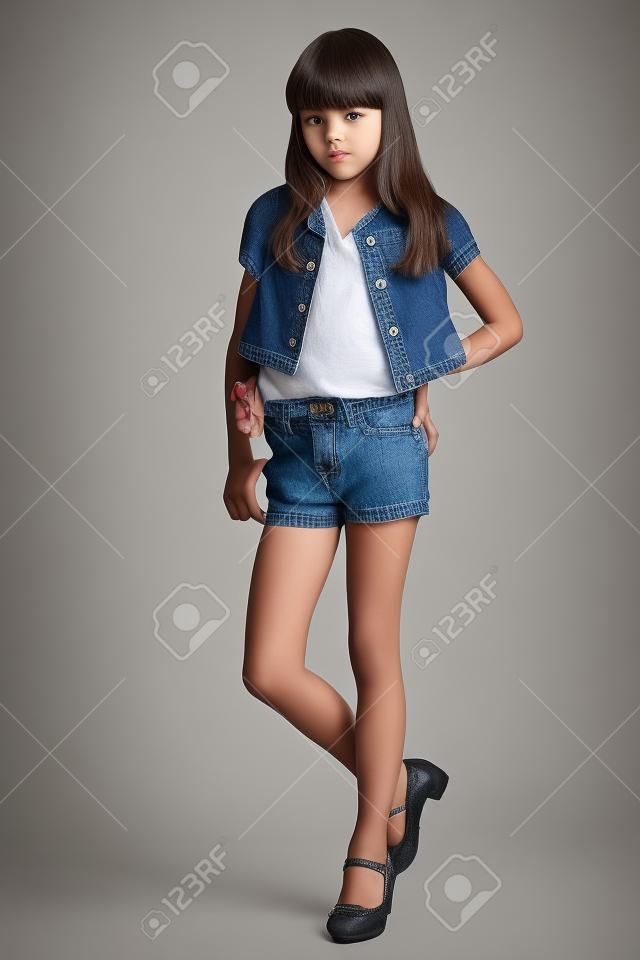 Piękna dziewczyna w dżinsowych szortach stoi na całej długości. Eleganckie atrakcyjne dziecko o smukłym ciele i długich nogawkach w rajstopach. Młoda uczennica ma 9 lat.