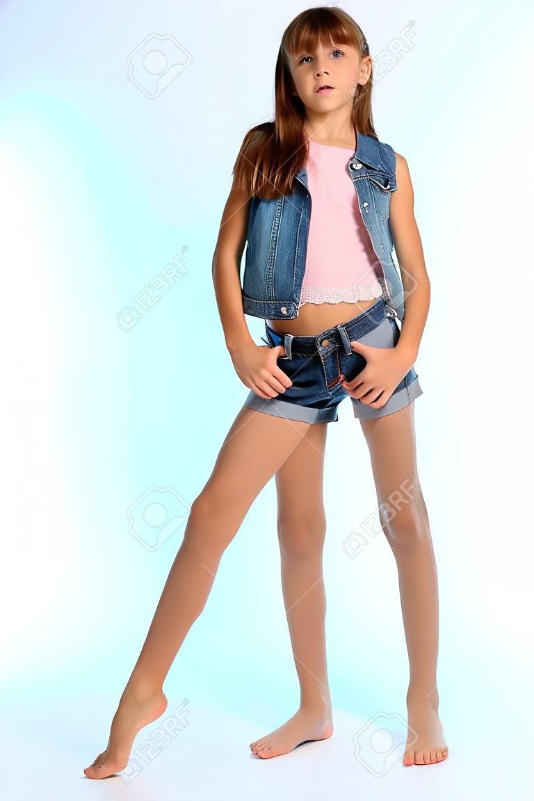 Menina bonita em um shorts denim está em pé em comprimento total. Criança atraente elegante com um corpo esbelto e pernas longas em meia-calça. A jovem estudante tem 9 anos de idade.