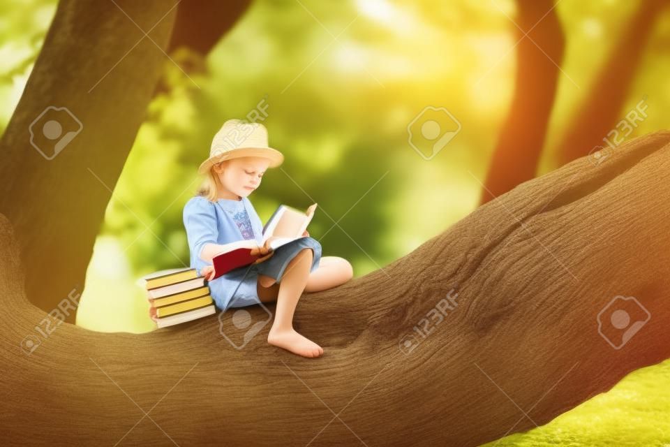 A cute little girl with blonde hair and a straw hat is reading a book in a big tree. Children and science.