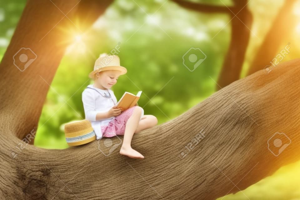 Ein süßes kleines Mädchen mit blonden Haaren und einem Strohhut liest ein Buch in einem großen Baum. Kinder und Wissenschaft.