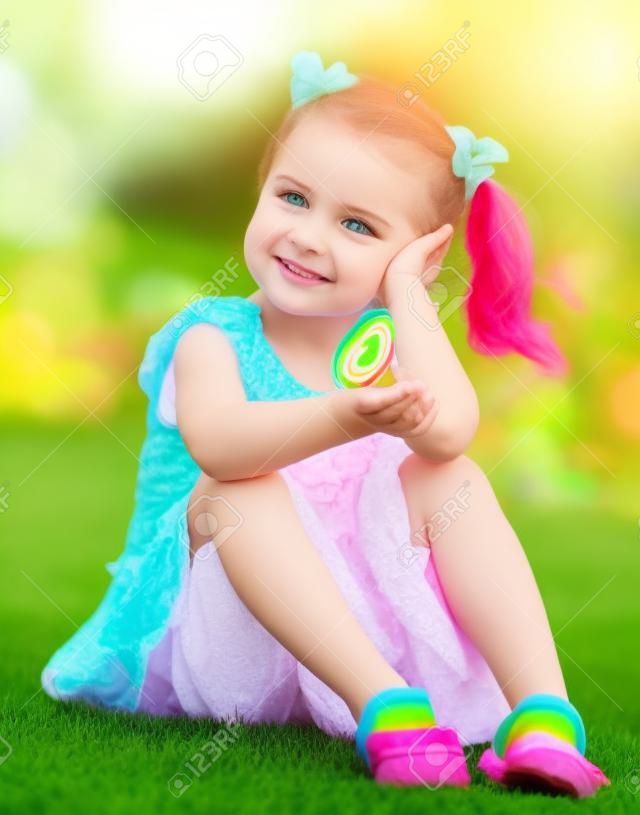 niña linda que se sienta en la hierba verde en el patio trasero y sosteniendo en la mano colorida del lollipop