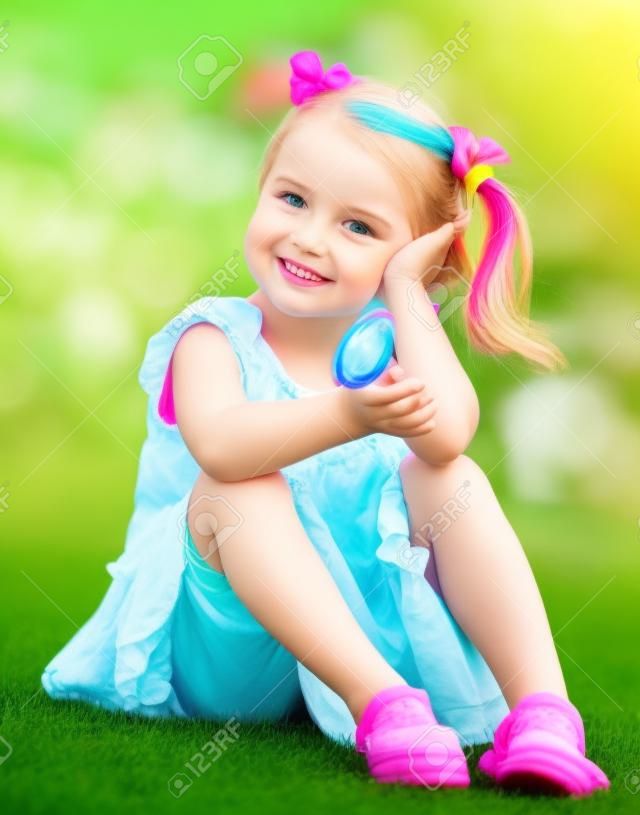 niña linda que se sienta en la hierba verde en el patio trasero y sosteniendo en la mano colorida del lollipop
