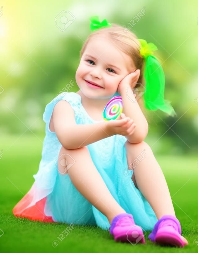  cute little girl sitting on green grass on backyard and holding in hand colorful lollipop