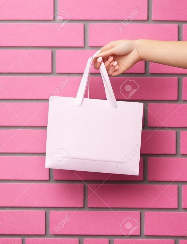 Sacchetto della spesa o pacchetto di carta di colore rosa-chiaro in mano femminile come presente o regalo di festa sul fondo del muro di mattoni, spazio della copia