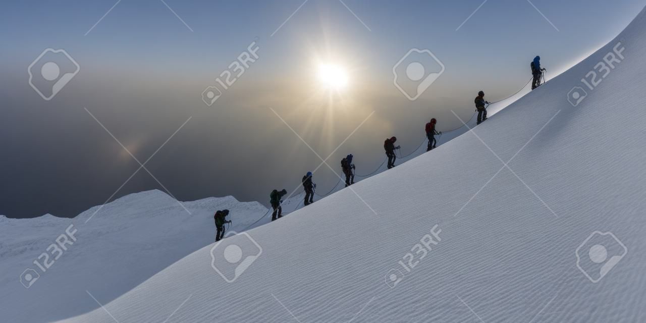 A roped party of experienced mountaineers climb the snowy side of a mountain to reach the summit. On the horizon the sun sets over the magical landscape.