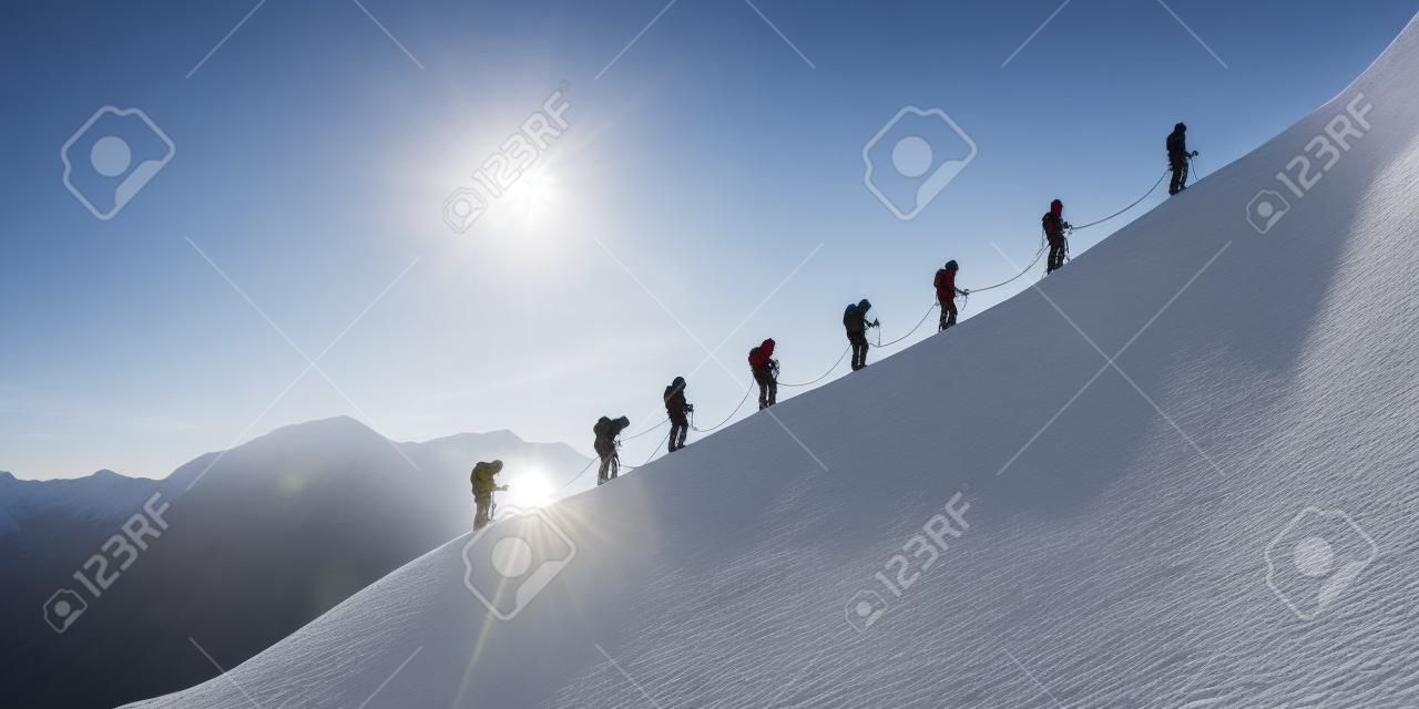 Um grupo de montanhistas experientes sobe o lado nevado de uma montanha para chegar ao cume. No horizonte, o sol se põe sobre a paisagem mágica.