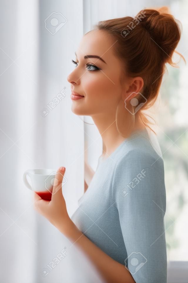 Portrait of the beautiful woman. She is drinking tea at the window