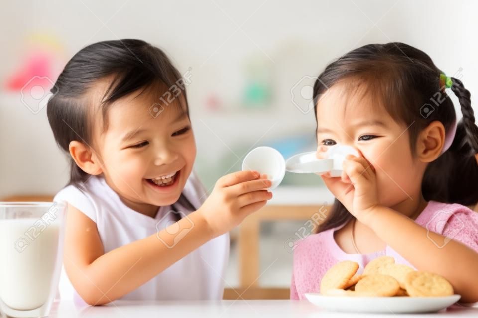 Zwei niedliche asiatische kleine Mädchen essen Kekse mit Milch für Frühstück zusammen mit Glück