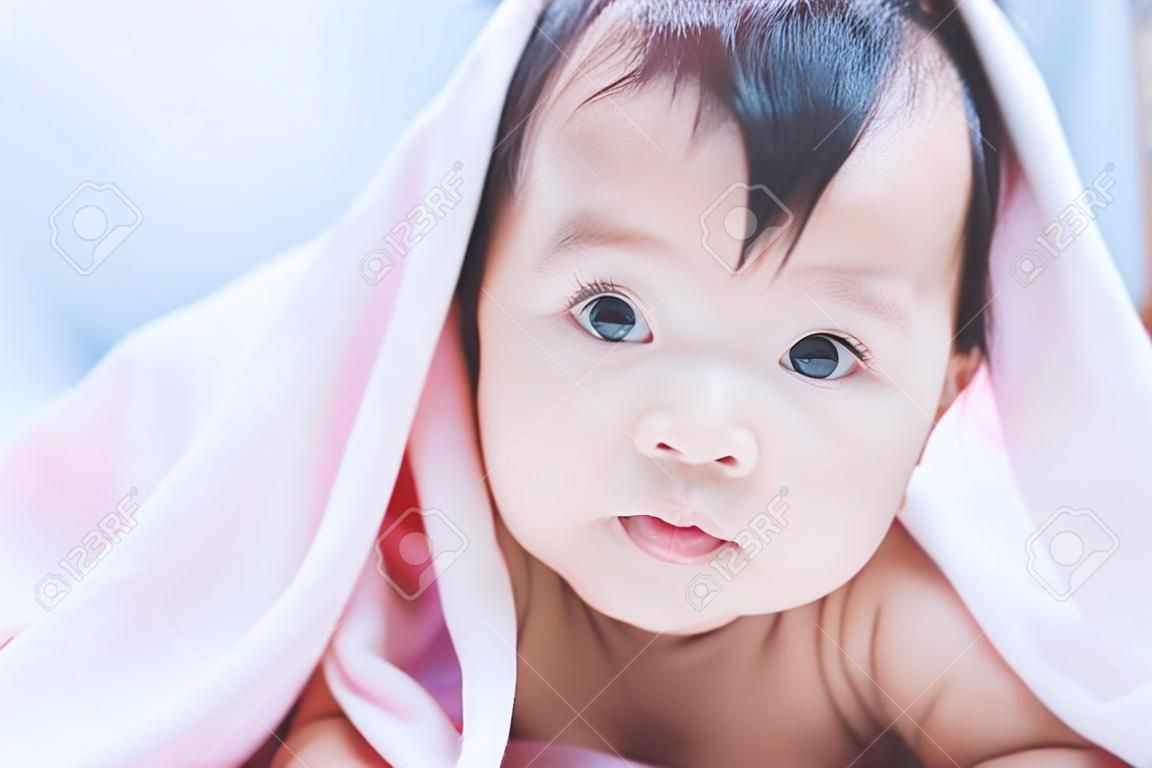 Cute asian baby girl smiling under pink blanket and lying on her stomach on the bed