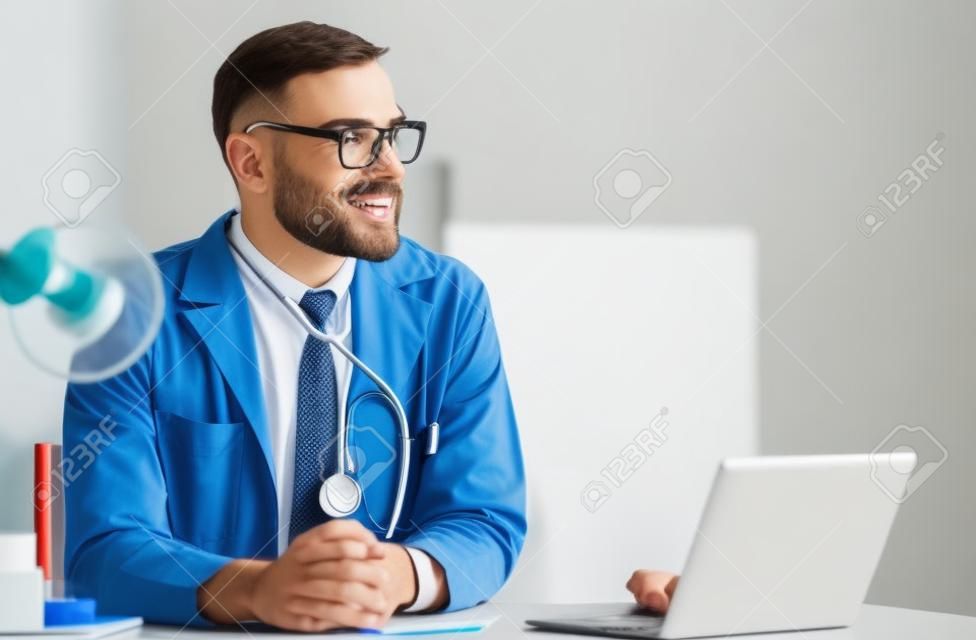 Happy male doctor at office desk in hospital working on laptop computer. Medical healthcare concept.
