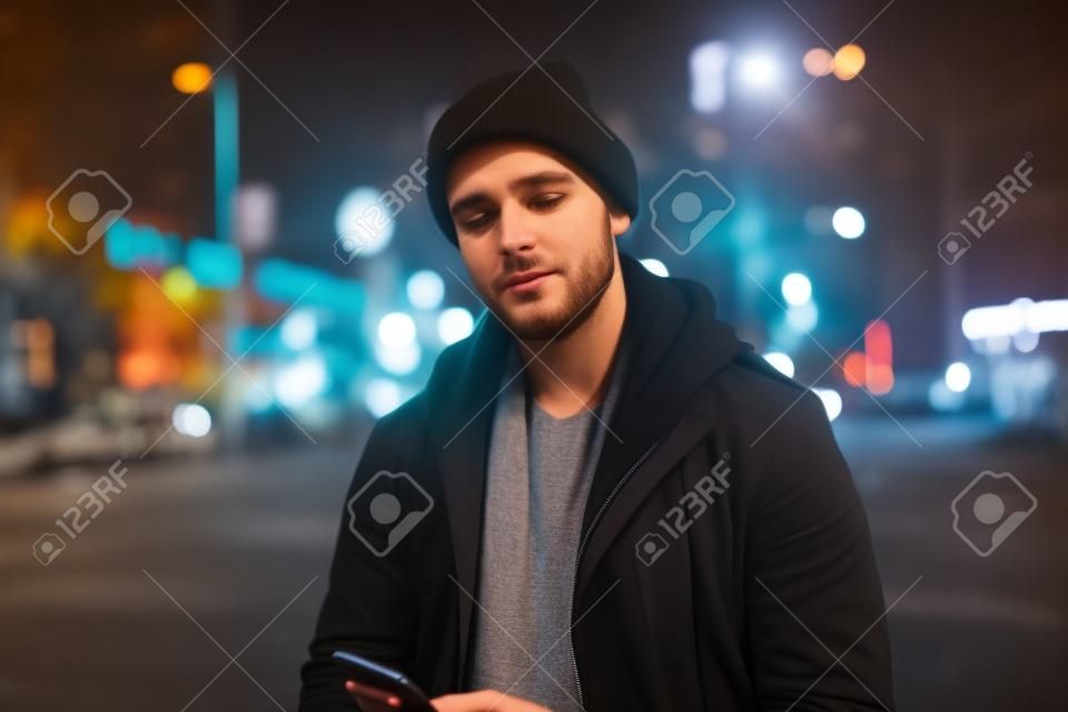 Wide angle shot of a young caucasian man trendy clothes using mobile phone with background of urban city street at night. Generative AI AIG18.