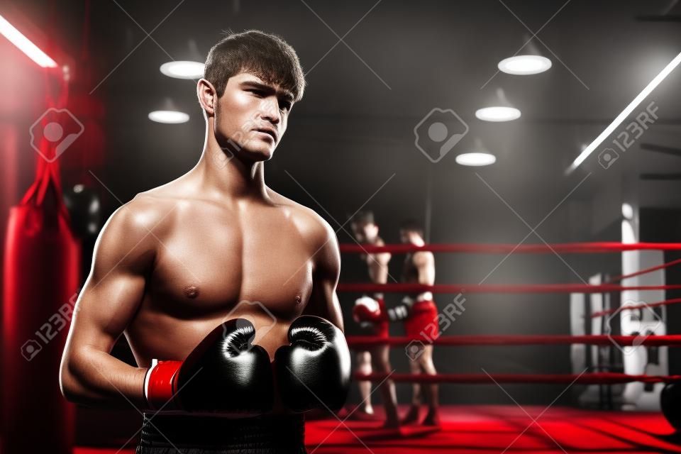 Boxing fighter shirtless posing, caucasian man boxer wearing red glove in defensive guard stance ready to fight and punch at gym with ring and boxing equipment in background. Impetus