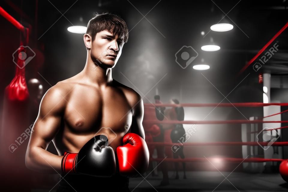Boxing fighter shirtless posing, caucasian man boxer wearing red glove in defensive guard stance ready to fight and punch at gym with ring and boxing equipment in background. Impetus