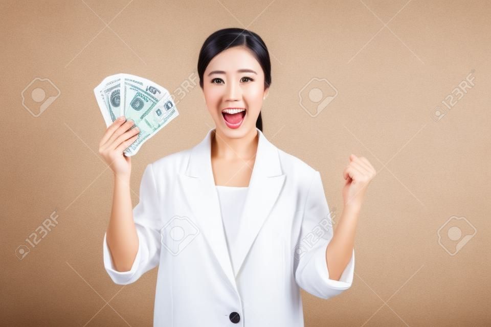 portrait of successful happy confident young asian business woman wearing white jacket holding cash money dollars standing over beige background. millionaire business, shopping concept.