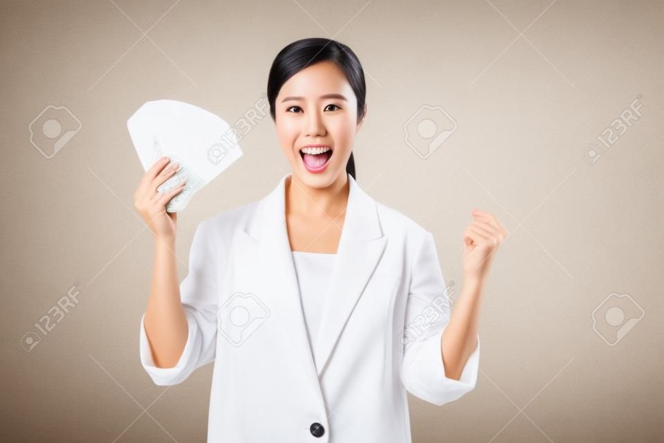 portrait of successful happy confident young asian business woman wearing white jacket holding cash money dollars standing over beige background. millionaire business, shopping concept.