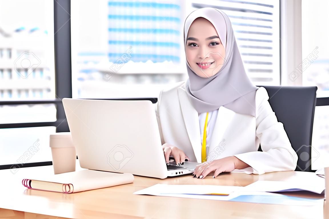 자신감 있는 아시아 이슬람(이슬람) 경제인 중역은 종교적인 베일을 입고 현대적인 사무실에서 일하고 카메라를 보고 미소를 짓고 있습니다. 문화의 다양성의 작업 개념입니다.