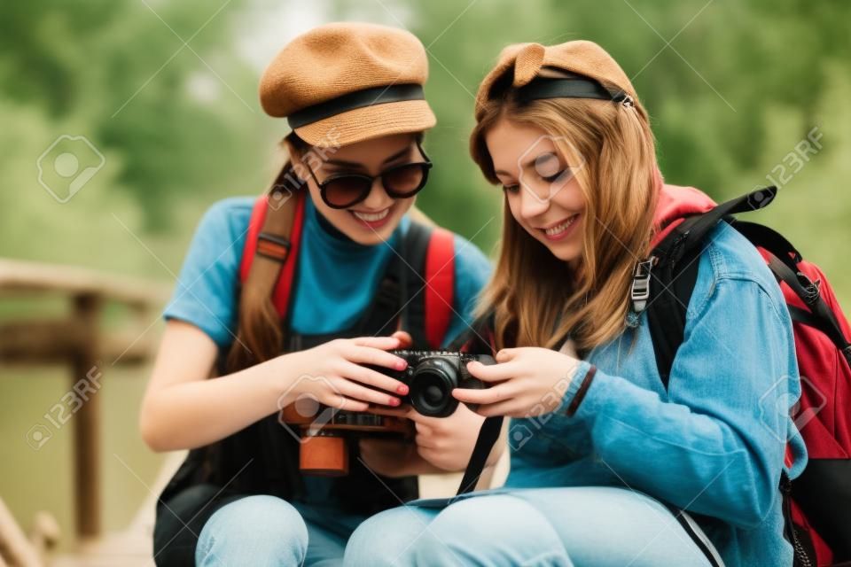 Duas meninas hipster legal da moda, amigos, na antiga ponte de madeira, e mochilas, segurando câmera vintage, emoções positivas.