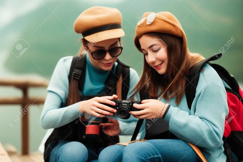 Dos modernas chicas hipster, amigas, en el viejo puente de madera y mochilas, sosteniendo una cámara vintage, emociones positivas.