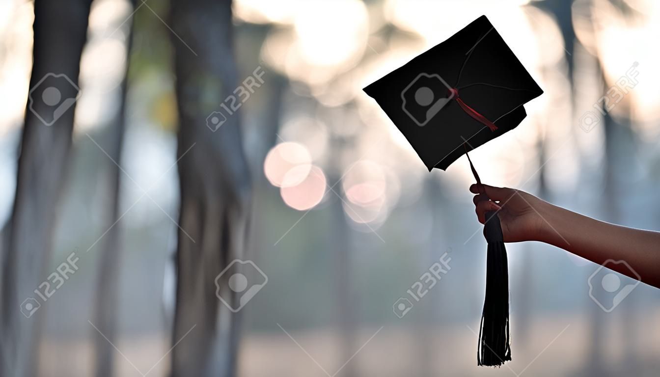 Mano de mujer graduada sosteniendo el gorro de graduación en su mano sobre fondo de bosque borroso. Concepto de celebración de graduación.