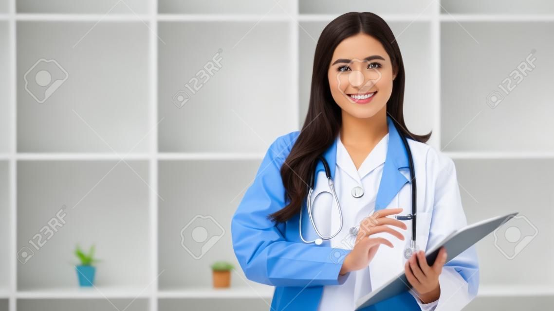 Portrait of female doctor wearing white coat with stethoscope holding clipboard and looking confidently to camera.