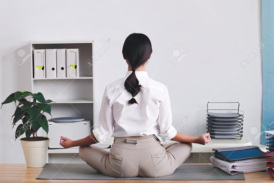 Vue arrière de la femme de bureau brune assise en position du lotus sur la table et méditant en silence