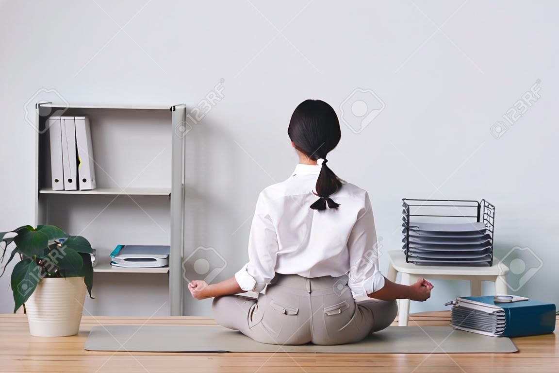 Vue arrière de la femme de bureau brune assise en position du lotus sur la table et méditant en silence