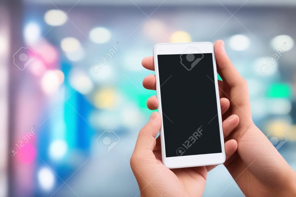 Man`s hand holding mobile smart phone with blank screen at blurred shopping mall background.