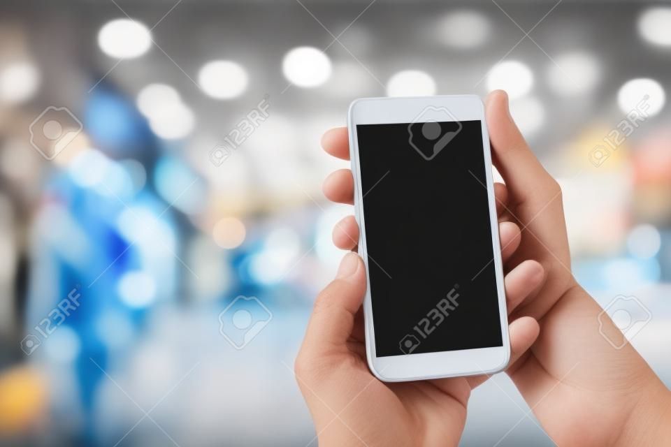 Manns Hand, die mobiles Smartphone mit leerem Bildschirm am unscharfen Einkaufszentrumhintergrund hält.