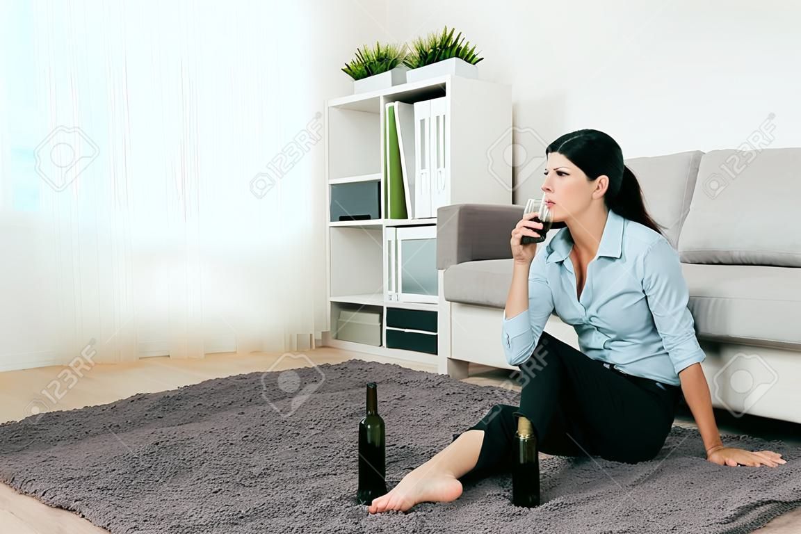 ビジネスウーマンマネージャーは仕事から悲しみを飲み去り、自宅のリビングルームの床に座っています