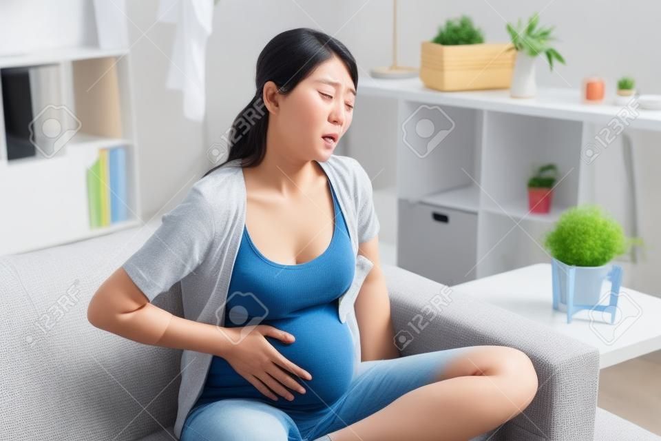 Jonge zwangere Aziatische vrouw lijden aan buikpijn thuis. ongemak Koreaanse toekomstige moeder met pijnlijke buik pijn baby groeien binnen lichaam. fronsen verdrietig ziek meisje zittend op de bank rust.