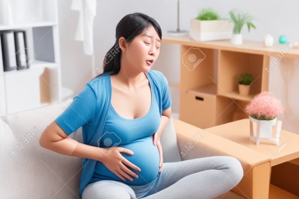 Jonge zwangere Aziatische vrouw lijden aan buikpijn thuis. ongemak Koreaanse toekomstige moeder met pijnlijke buik pijn baby groeien binnen lichaam. fronsen verdrietig ziek meisje zittend op de bank rust.