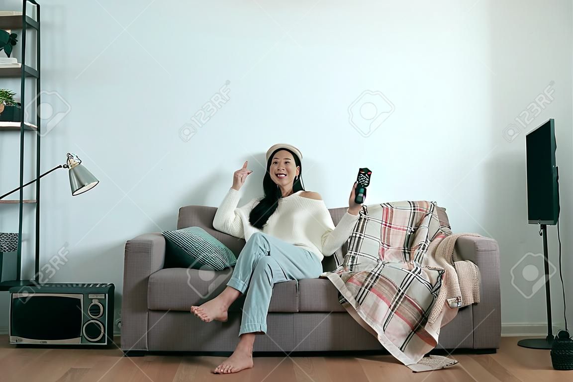 Unbeschwerte asiatische Frau, die zu Hause auf dem Sofa in der modernen Winterwohnung fernsieht. Junges lachendes lässiges Mädchen, das Fernsehsendung mit Fernbedienung genießt. Komfortleben Lifestyle Exemplar copy