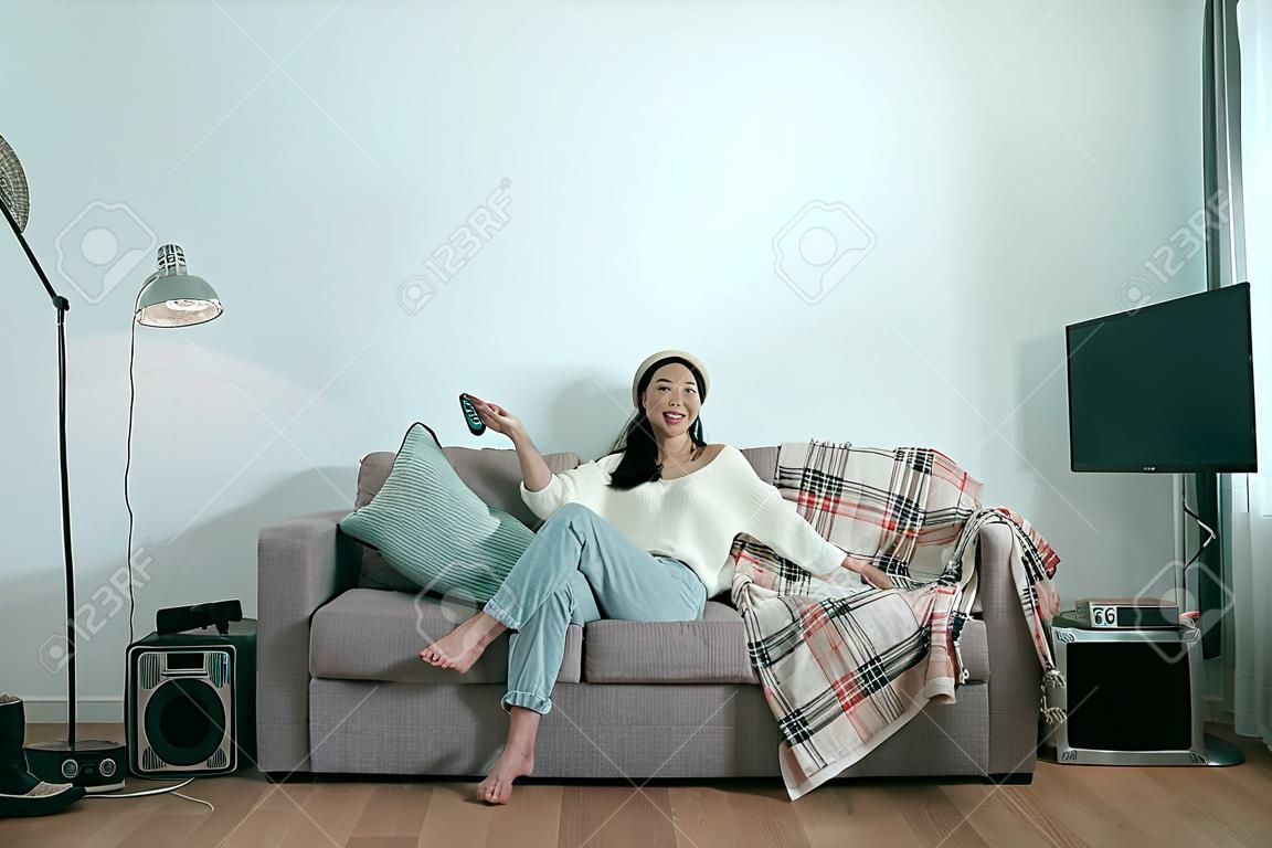 carefree asian woman watching TV at home sitting on sofa in modern winter apartment. Young laughing casual girl enjoying television show holding control remote. Comfort living lifestyle copyspace