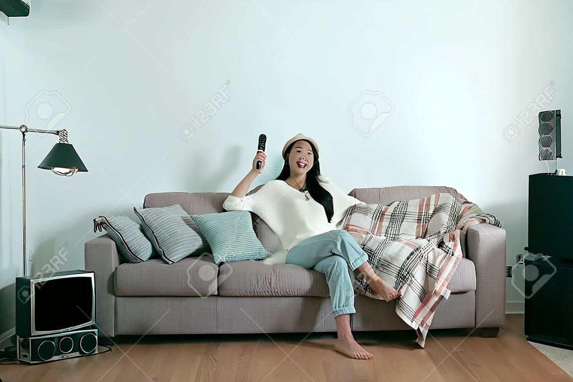 zorgeloos Aziatische vrouw kijken tv thuis zitten op de bank in moderne winter appartement. Jonge lachende casual meisje genieten tv-show holding control afstandsbediening. Comfort levende levensstijl copyspace