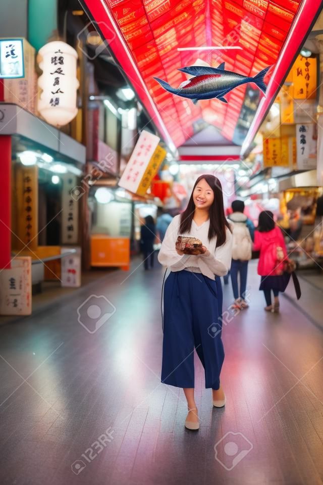 vertical full length of curious asian girl holding camera looking at different stalls while walking along the street in kuromon ichiba market with fish decoration above head in Osaka japan