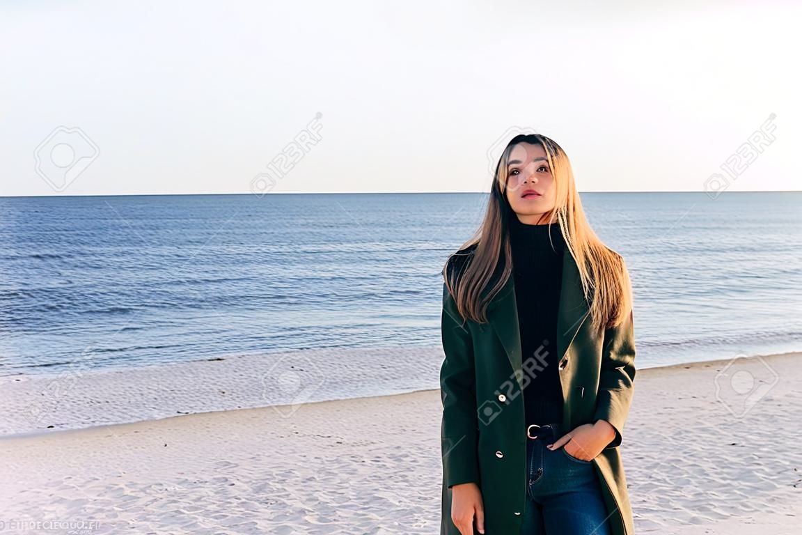Young woman wearing stylish coat, black turtleneck and jeans posing on the autumn beach. Portrait of girl looking at camera at sunny day by the sea.
