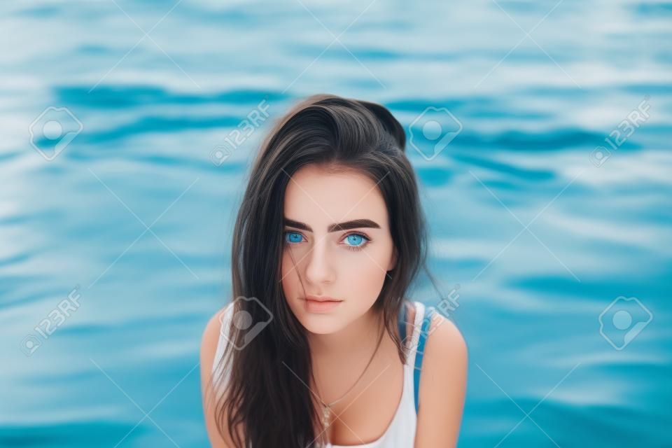 Portrait of cute girl with long dark hair, big eyebrows and blue eyes, resting near water. Young woman wearing white top sitting on the background of sea surface looking at camera.