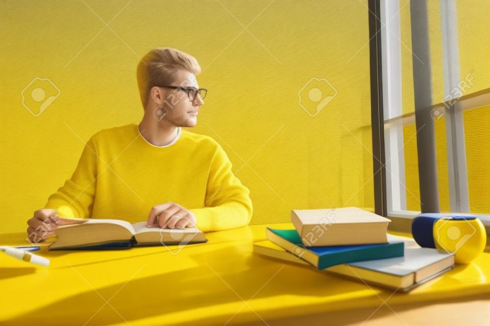 Blonde student in glasses wearing yellow pullover looking to the window while doing his homework at the class