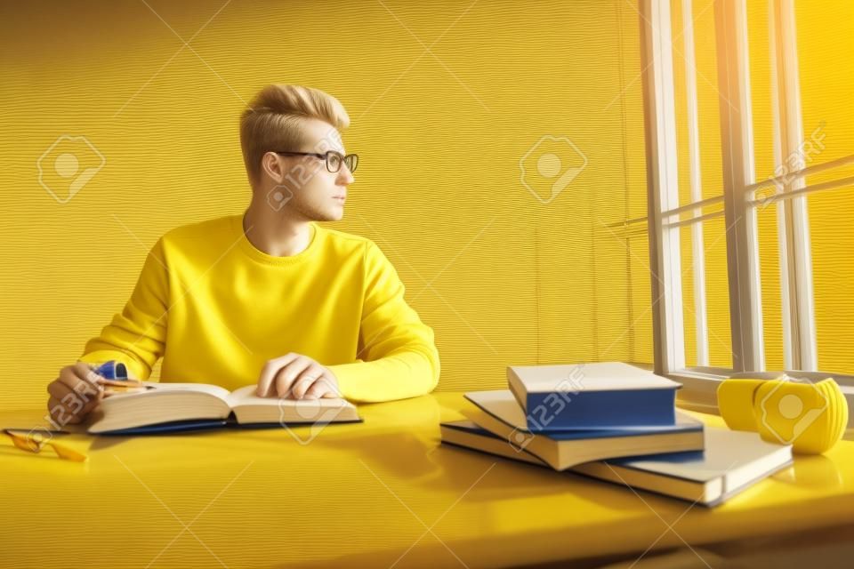 Estudante loiro em óculos usando pulôver amarelo olhando para a janela enquanto faz sua lição de casa na classe