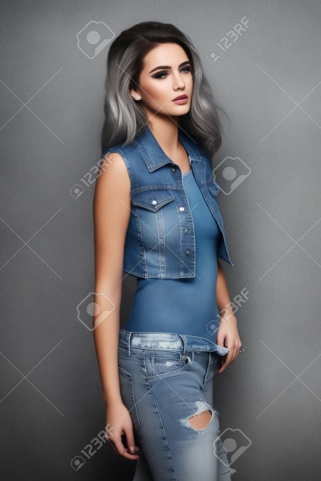 Mujer joven imponente en ropa de jeans que presenta sobre fondo gris. estilo denim. Captura de moda.
