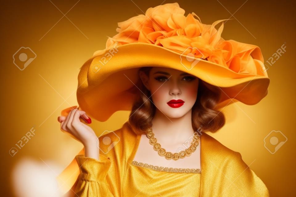 Portrait of a gorgeous young woman in elegant broad-brimmed hat and luxurious dress over golden background. Vintage style. Beauty, fashion.