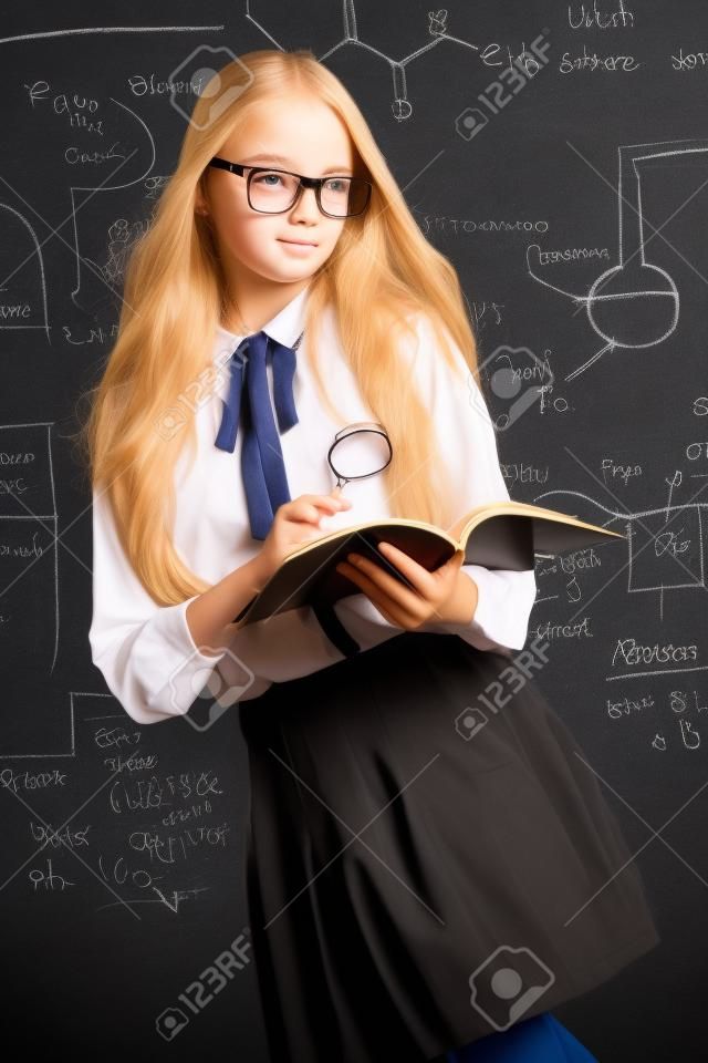 Muchacha linda del estudiante con el pelo rubio largo que presenta en uniforme escolar y vidrios sobre fondo de la pizarra.
