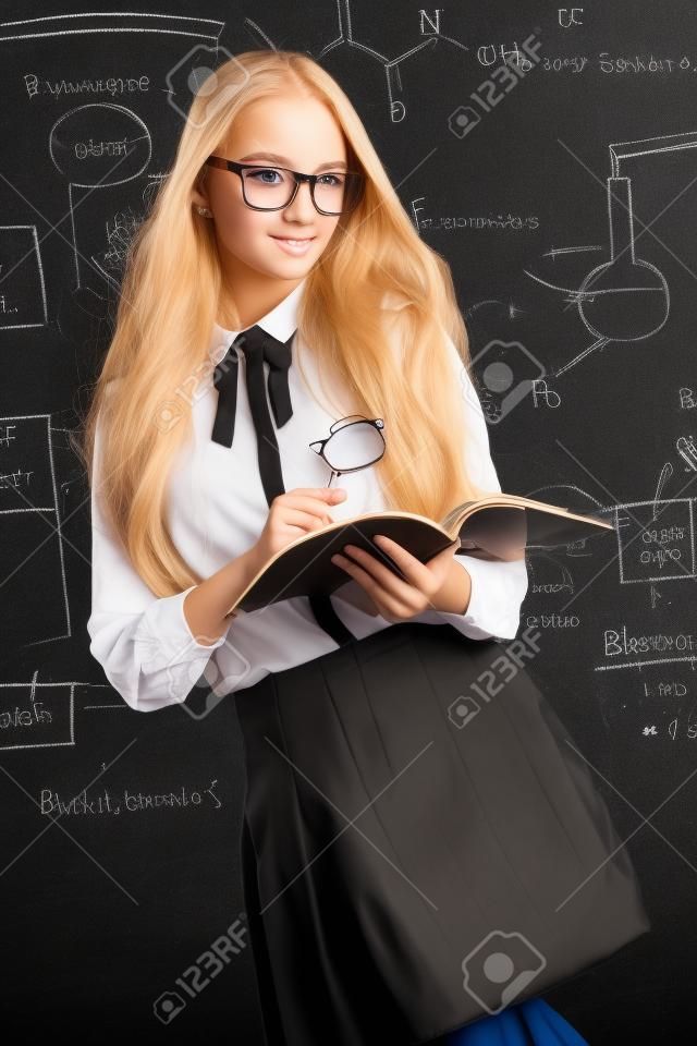 Muchacha linda del estudiante con el pelo rubio largo que presenta en uniforme escolar y vidrios sobre fondo de la pizarra.