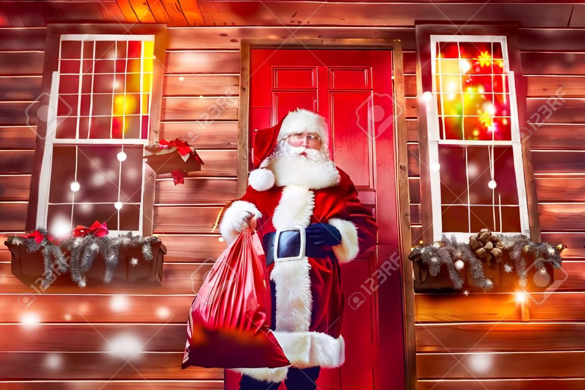 Portrait du bon vieux père Noël tenant un sac avec des cadeaux sur le porche de sa maison décorée. Maison du Père Noël. Concept de Noël et du nouvel an.