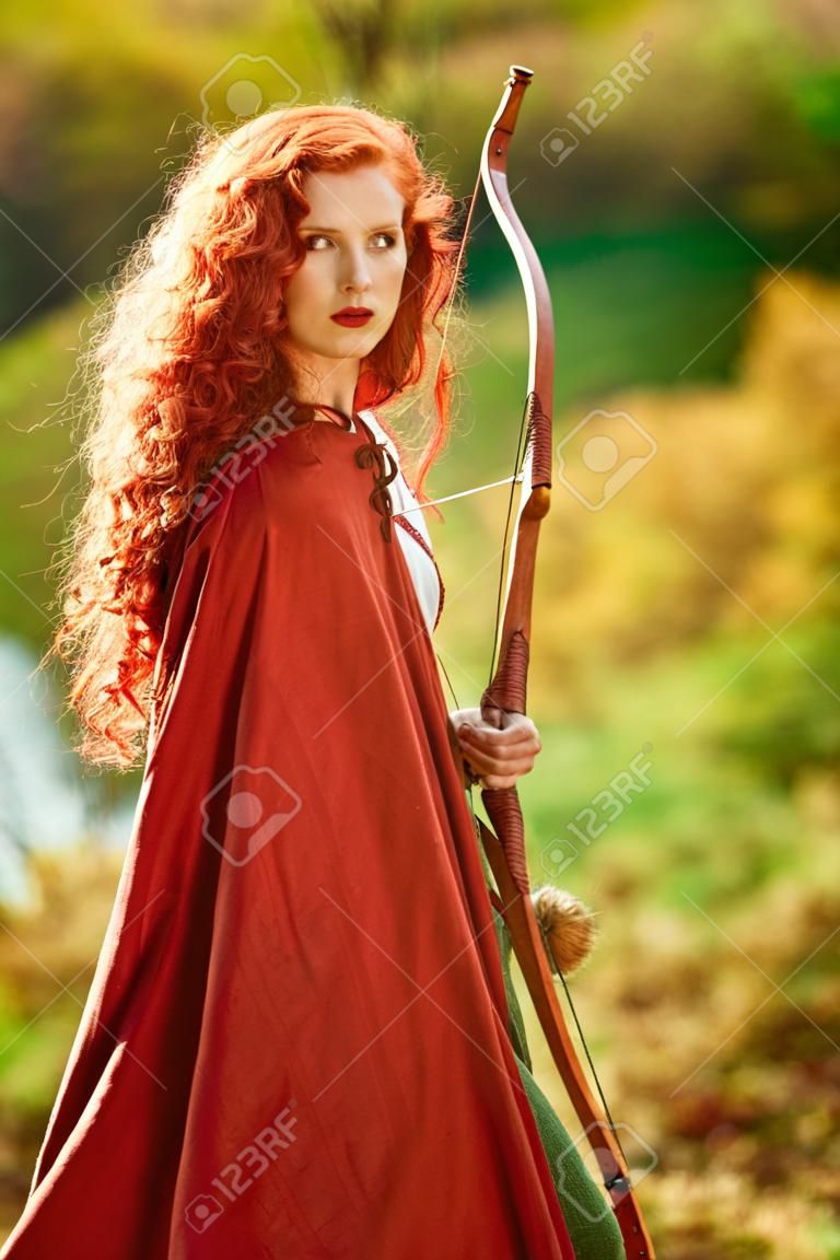 Historische reconstructie. Prachtige jonge vrouw boogschutter met prachtige lange rode haren in een historische keltische jurk staat aan de rand van het bos.