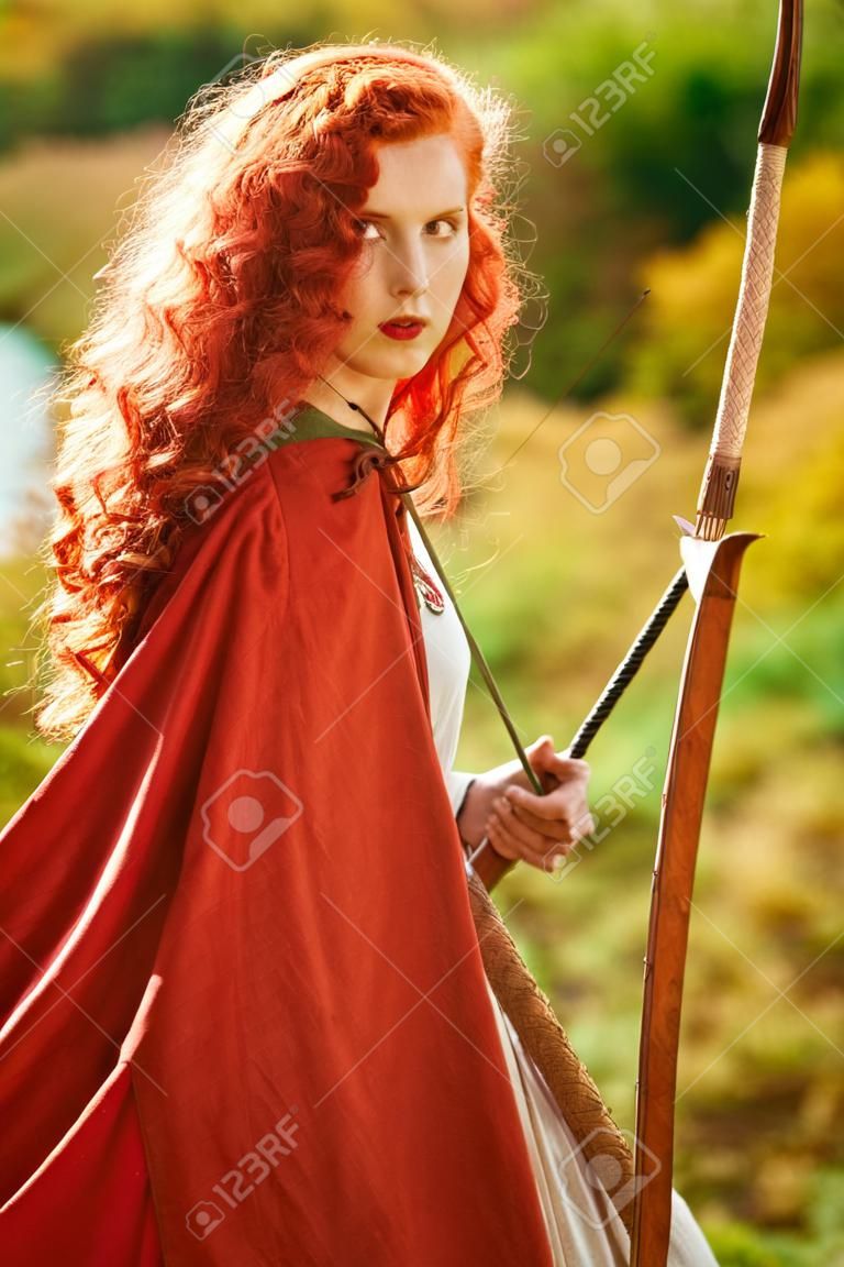 Historische reconstructie. Prachtige jonge vrouw boogschutter met prachtige lange rode haren in een historische keltische jurk staat aan de rand van het bos.